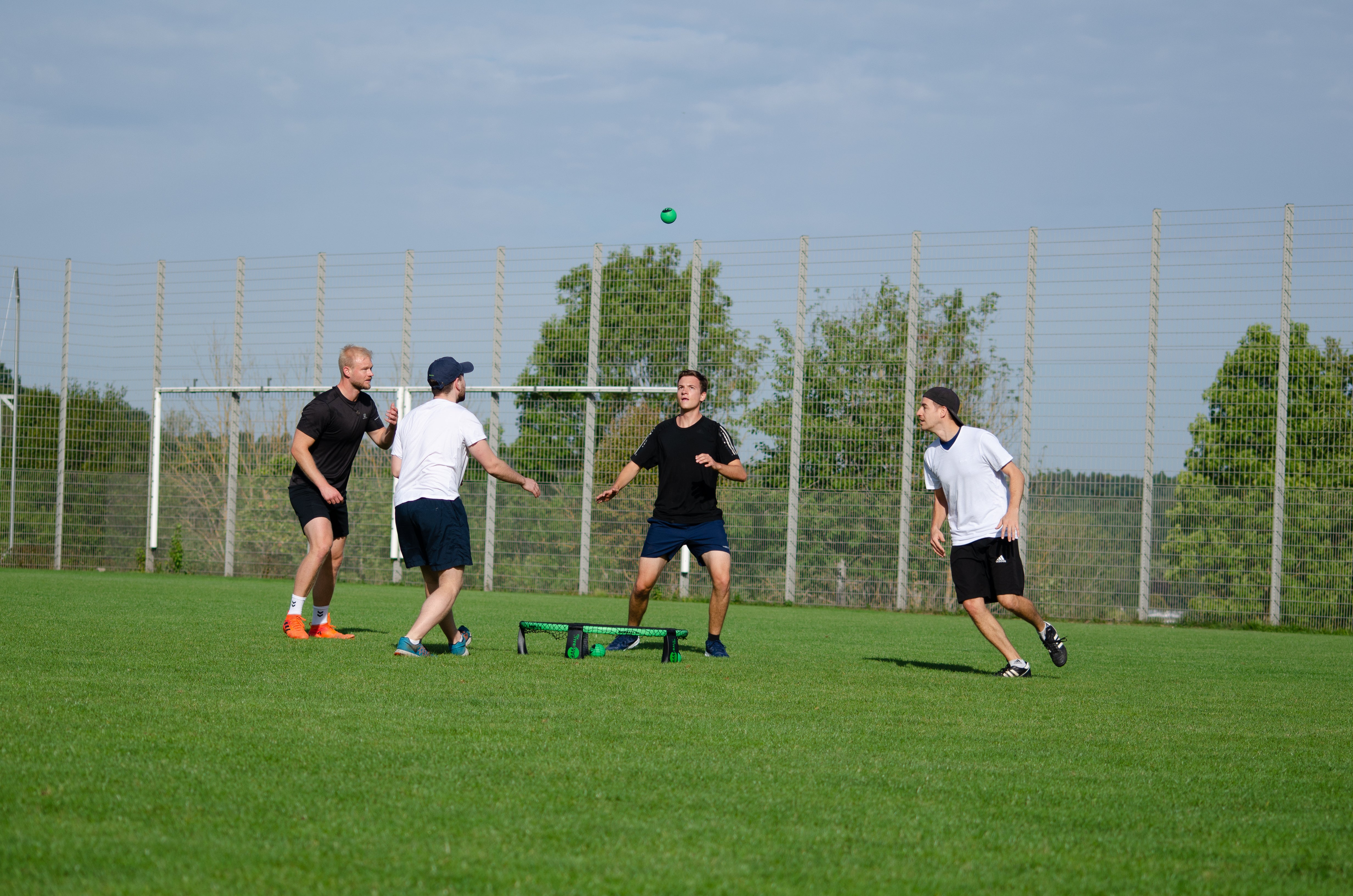 Vier Roundnet Spieler auf Gras, ein Sportler ist kurz davor, den Ball aufs Trampolin zu schlagen
