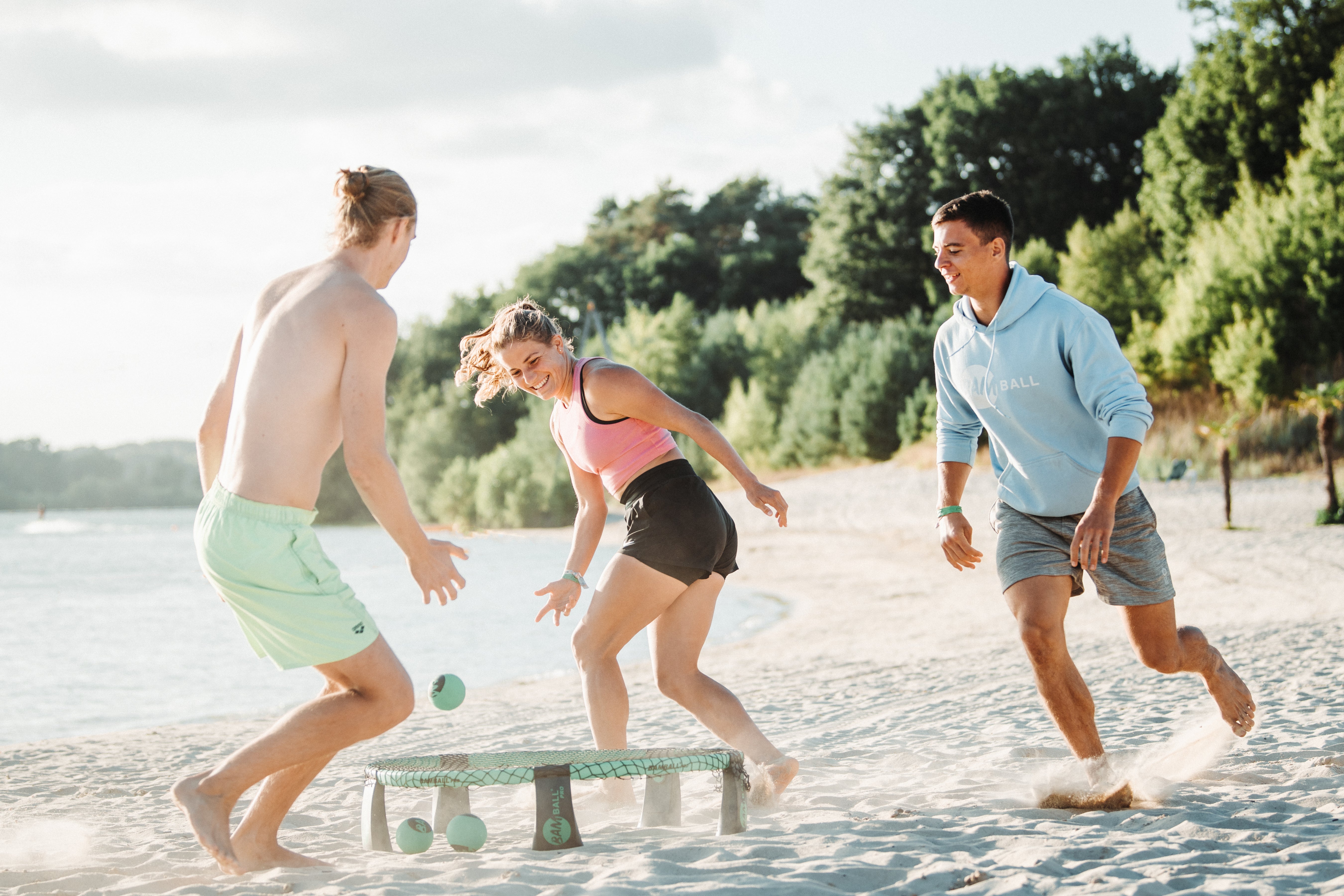 Drei Personen spielen den Roundnet-Sport am Strand, eine Spielerin spielt den Ball aufs Trampolin